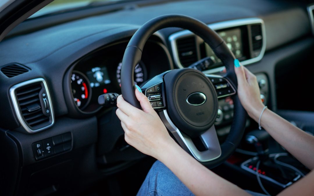 Person with turquoise nails driving a car, hands on the steering wheel, dashboard and speedometer visible in the background.