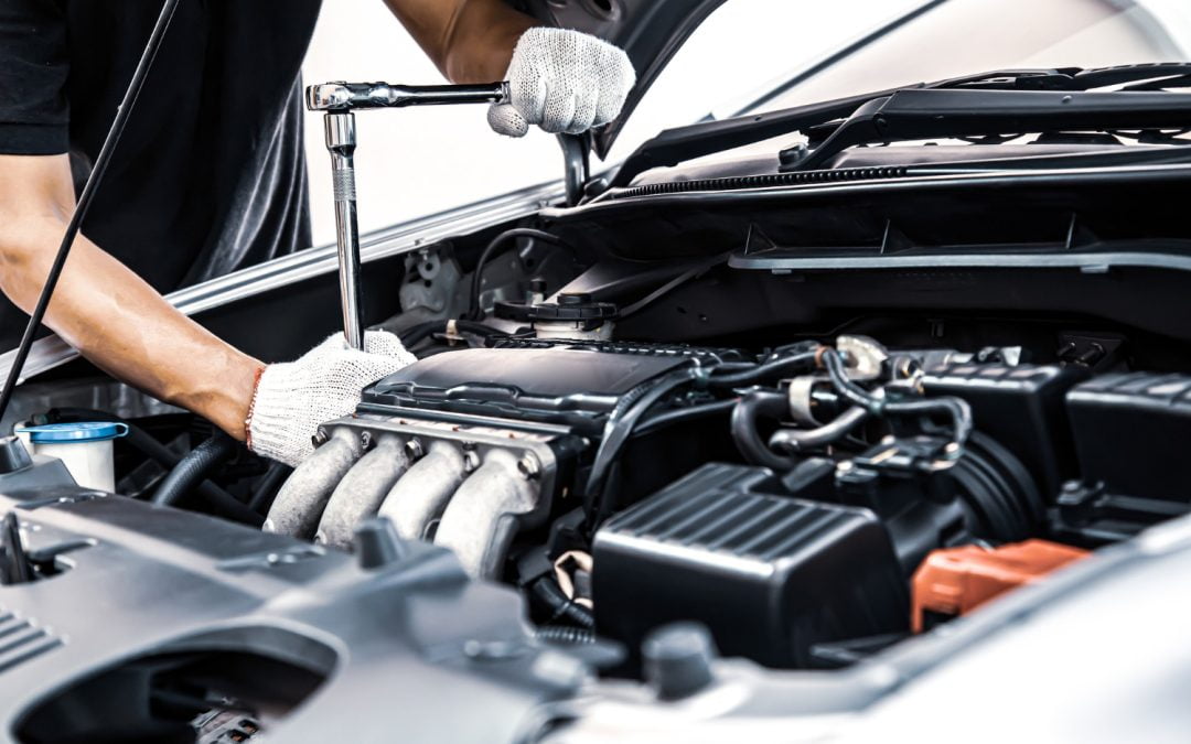 A mechanic wearing gloves uses a wrench to perform car maintenance under the open hood.