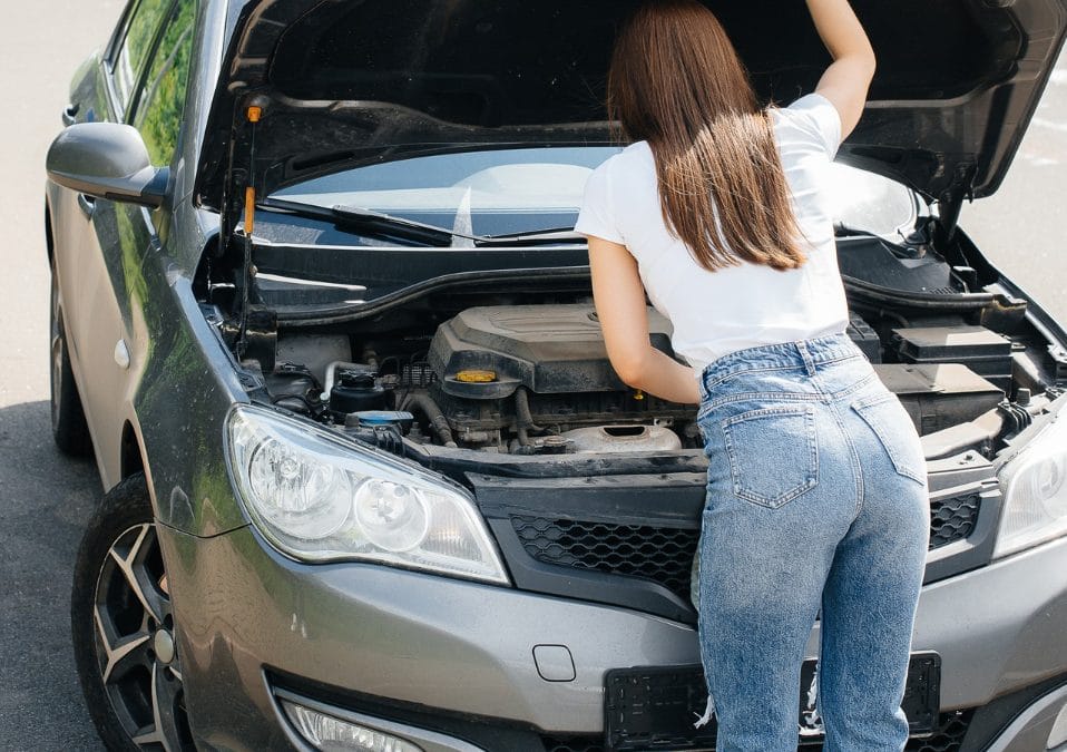 A woman stands by an open car hood on the roadside, looking at the engine.