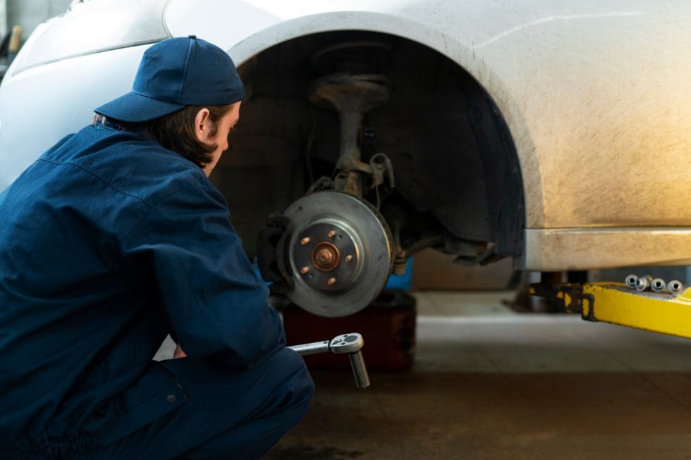 Mechanic in blue uniform inspecting a car’s exposed wheel hub with tools in hand at an auto shop.