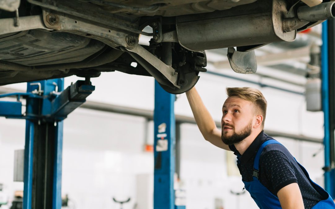 A mechanic in blue overalls inspects the underside of a car raised on a lift in a garage.