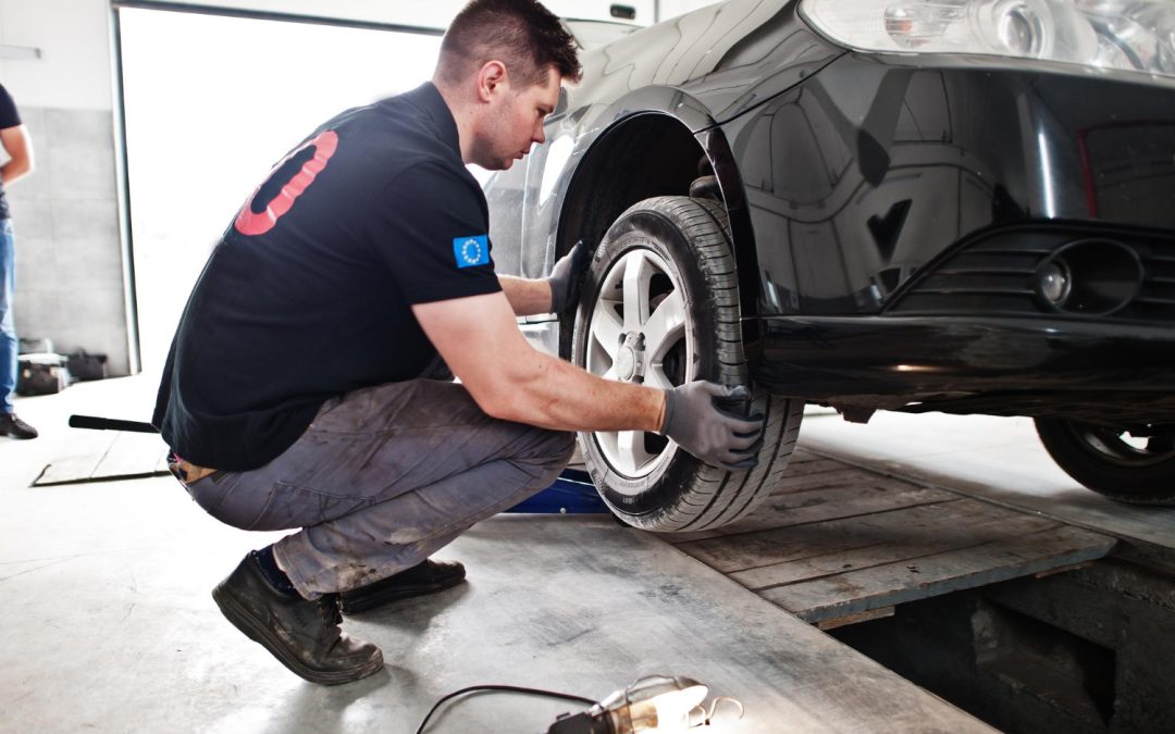 Mechanic in gloves squats, checking the front tire of a black car in a garage with tools nearby.